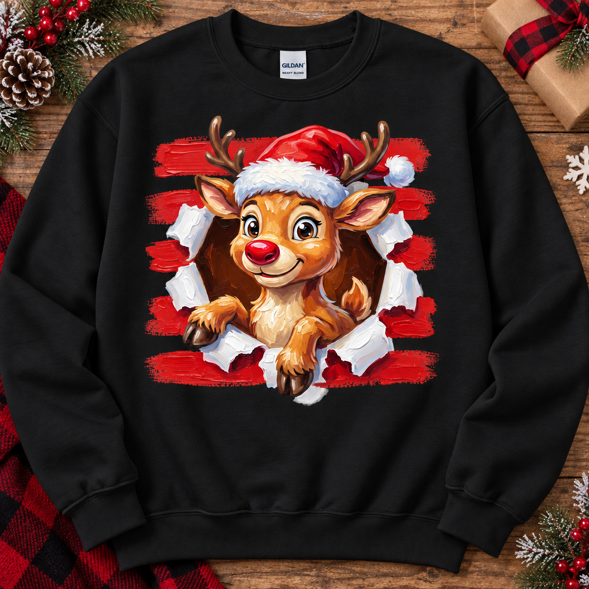 A black crewneck sweatshirt featuring a graphic of a cute cartoon reindeer wearing a Santa hat, bursting through torn red and white paper. The sweatshirt is displayed flat on a rustic wooden background decorated with Christmas pine branches, a gift box, and a red plaid scarf.