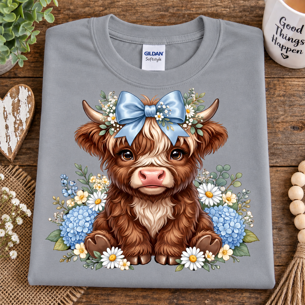 A folded sport gray Gildan Softee t-shirt with a cute cartoon Highland cow graphic, styled on a rustic wooden background with decorative props.