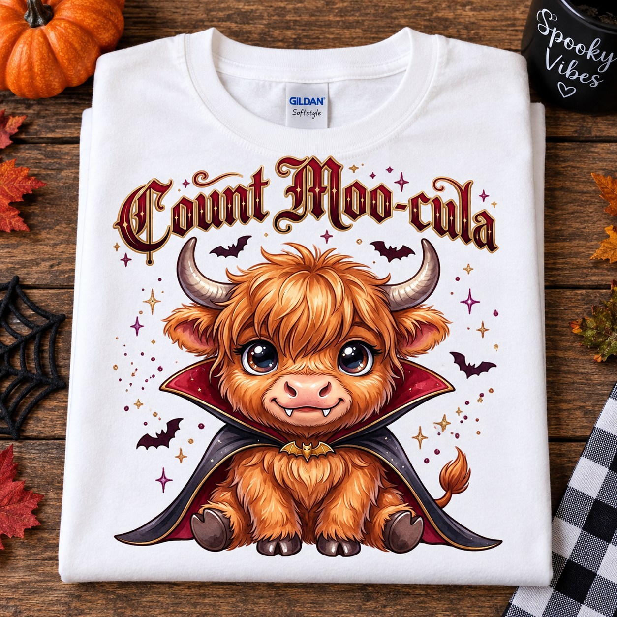 A folded white Gildan Softstyle T-shirt with a cute cartoon Highland cow dressed as a vampire, surrounded by bats, stars, and the text 'Count Moo cula,' styled on a wooden surface with autumn props.