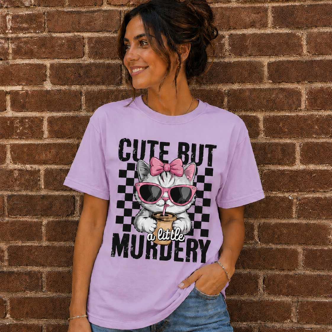 Woman wearing a Orchid graphic t-shirt with a cute kitten wearing pink sunglasses and the phrase “Cute But a Little Murdery” standing in front of a brick wall.