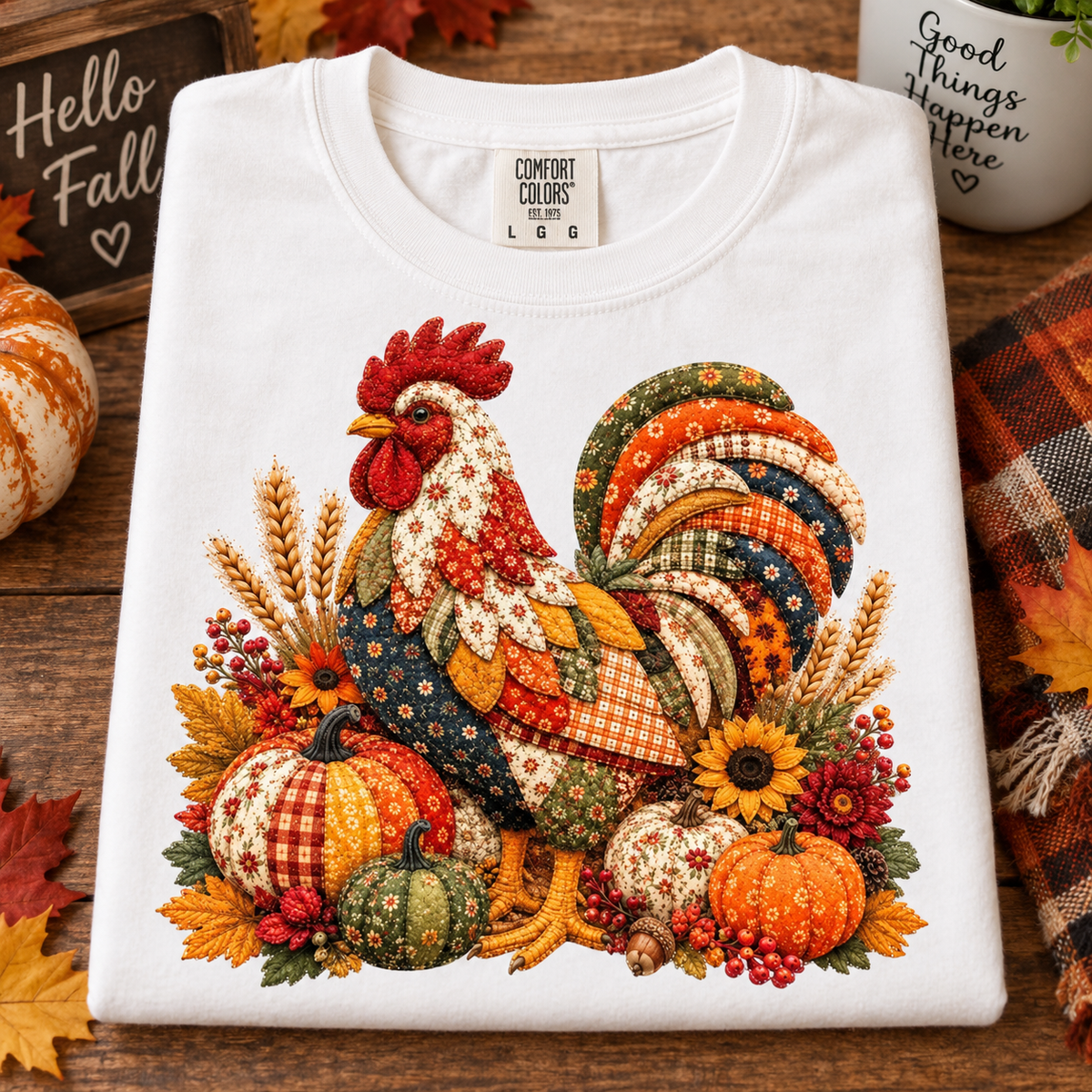 A folded white crewneck sweatshirt with a colorful autumn-themed rooster patchwork graphic, styled on a rustic wooden surface with fall decorations.