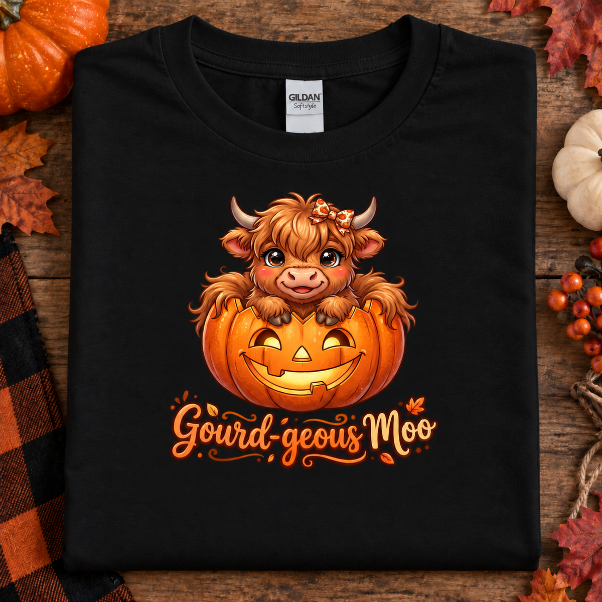 A black Gildan Comfort T-shirt laid flat on a rustic wooden surface, decorated with autumn props. The shirt features a cartoon illustration of a smiling Highland cow sitting inside a carved orange pumpkin, with the text 'Gourd-geous Moo' written below in orange script.