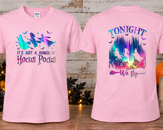 A light pink short-sleeve graphic T-shirts shown front and back on a styled tabletop setup. The front has a witch silhouette graphic with the text 'IT'S JUST A BUNCH OF Hocus Pocus.
