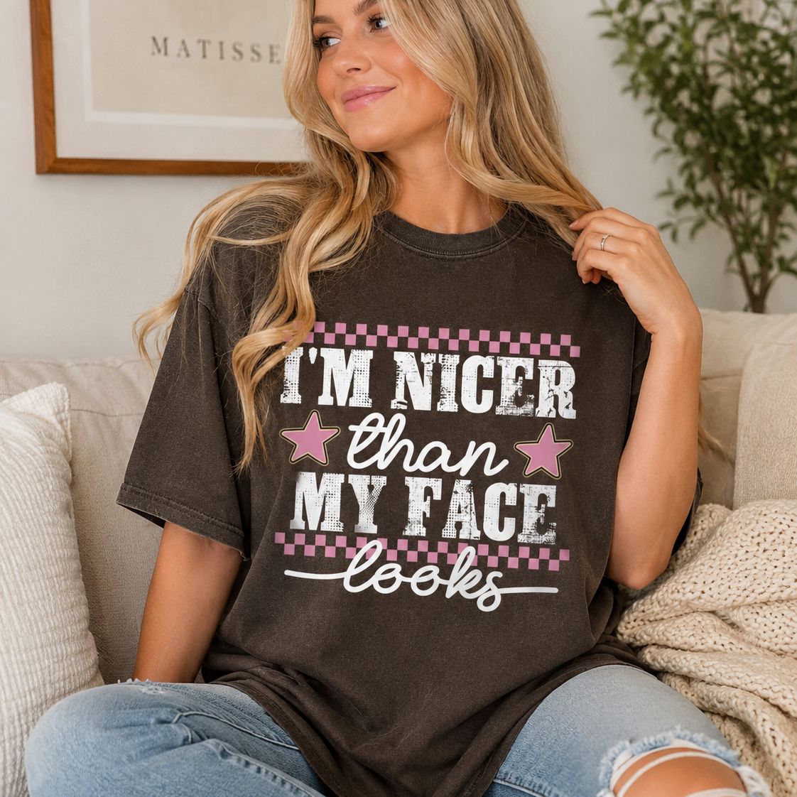 Woman with long blonde hair sitting on a couch wearing a espresso graphic t-shirt that says “I’m Nicer Than My Face Looks”