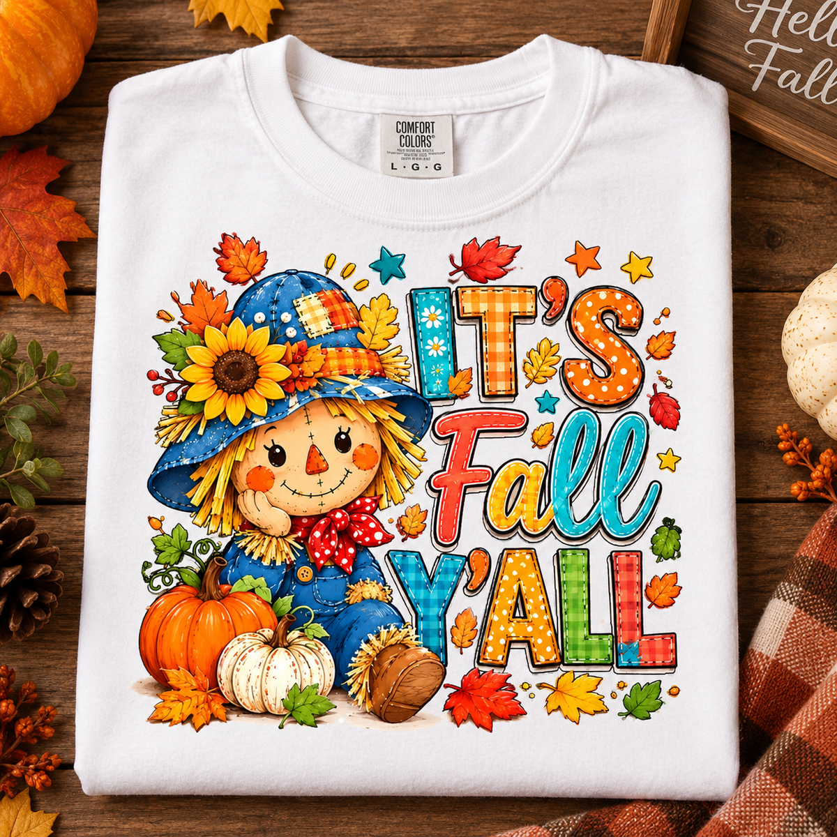 A folded white graphic T-shirt displayed on a rustic wooden surface with autumn props. The shirt features a colorful fall-themed design with a cartoon scarecrow, pumpkins, leaves, stars, and the text “IT’S Fall Y’ALL.