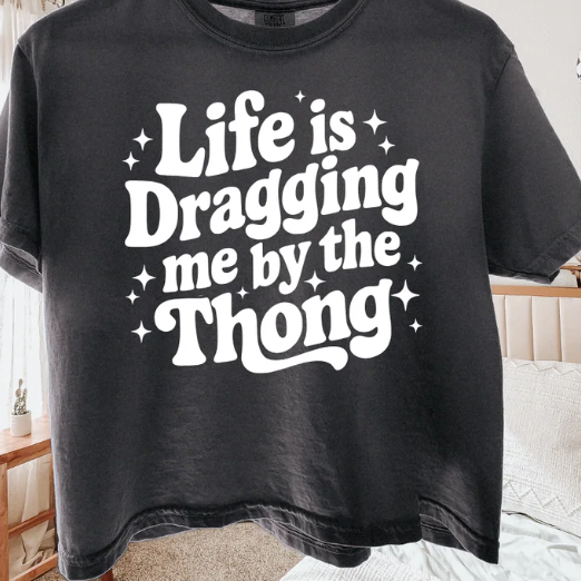 Life Is Dragging Me By the Thong graphic t-shirt with bold, funny lettering.