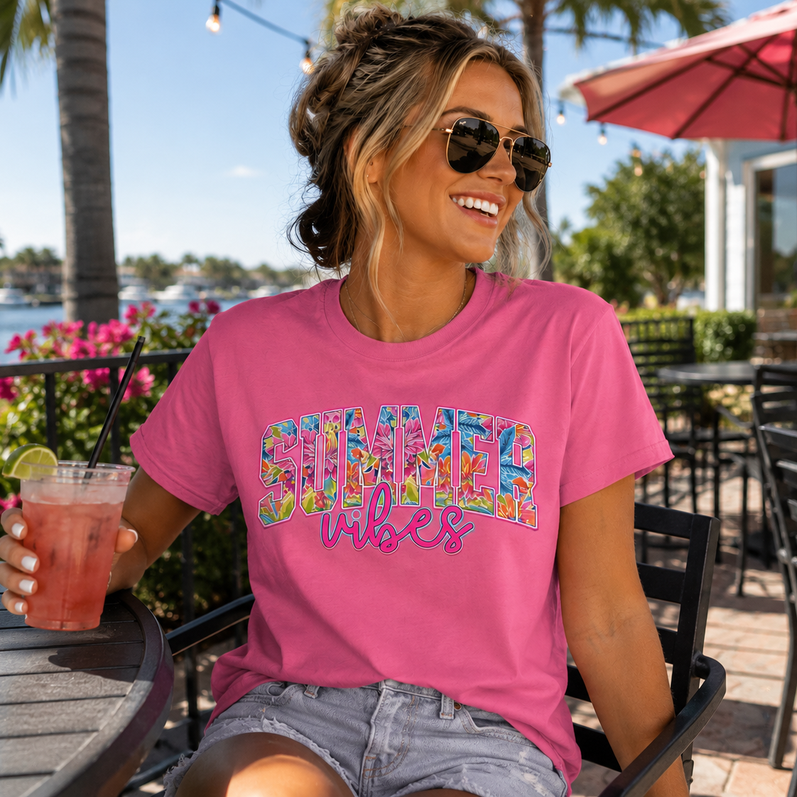 Woman wearing a azalea(pink) 'Summer Vibes' t-shirt outdoors with a drink.