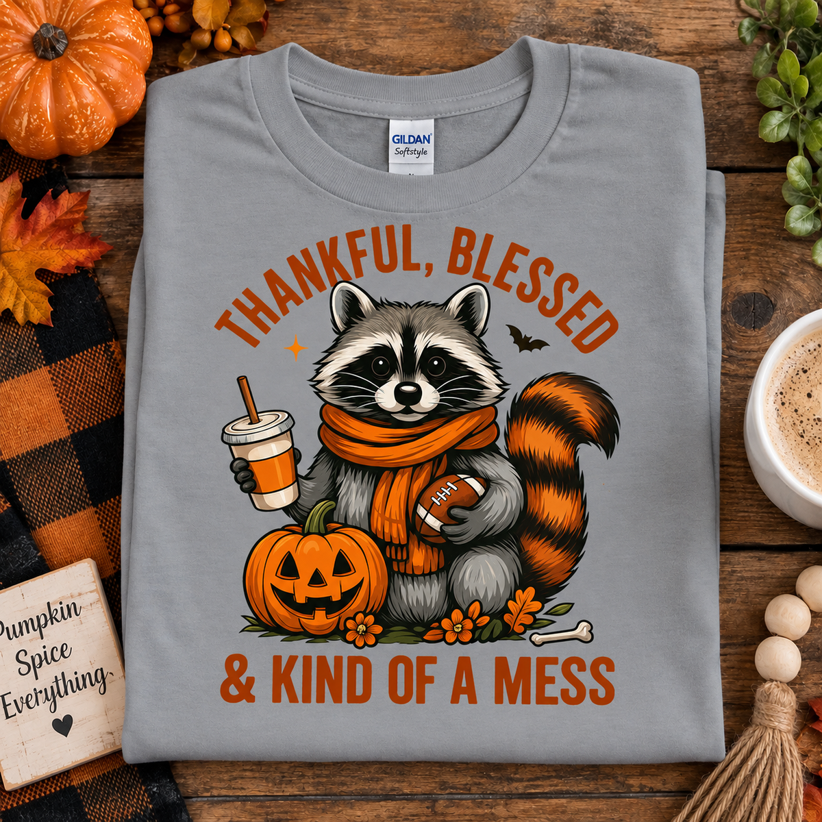 A folded sport gray Gildan Softstyle t-shirt with a cartoon raccoon graphic holding a football and pumpkin, surrounded by autumn props on a wooden surface.