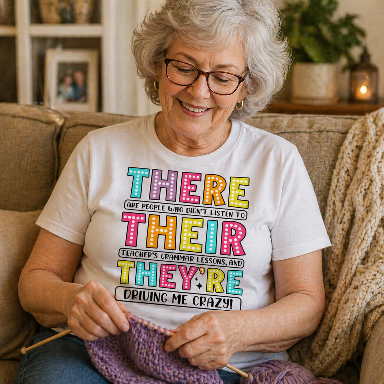 Senior woman sitting on a couch knitting, wearing a white t-shirt with colorful text.