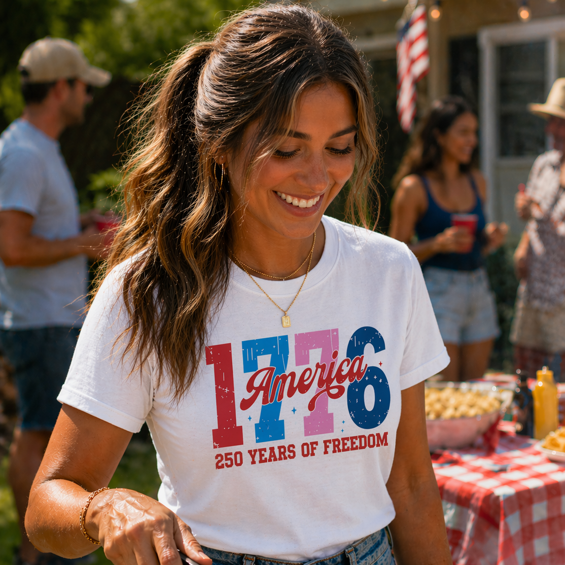 Woman wearing a white t-shirt with '1776 America 250 Years of Freedom' design, standing outdoors with people and a table in the background.