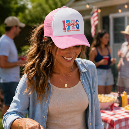 Woman grilling outdoors wearing a pink cap with '1776' text, surrounded by people at a casual outdoor event.