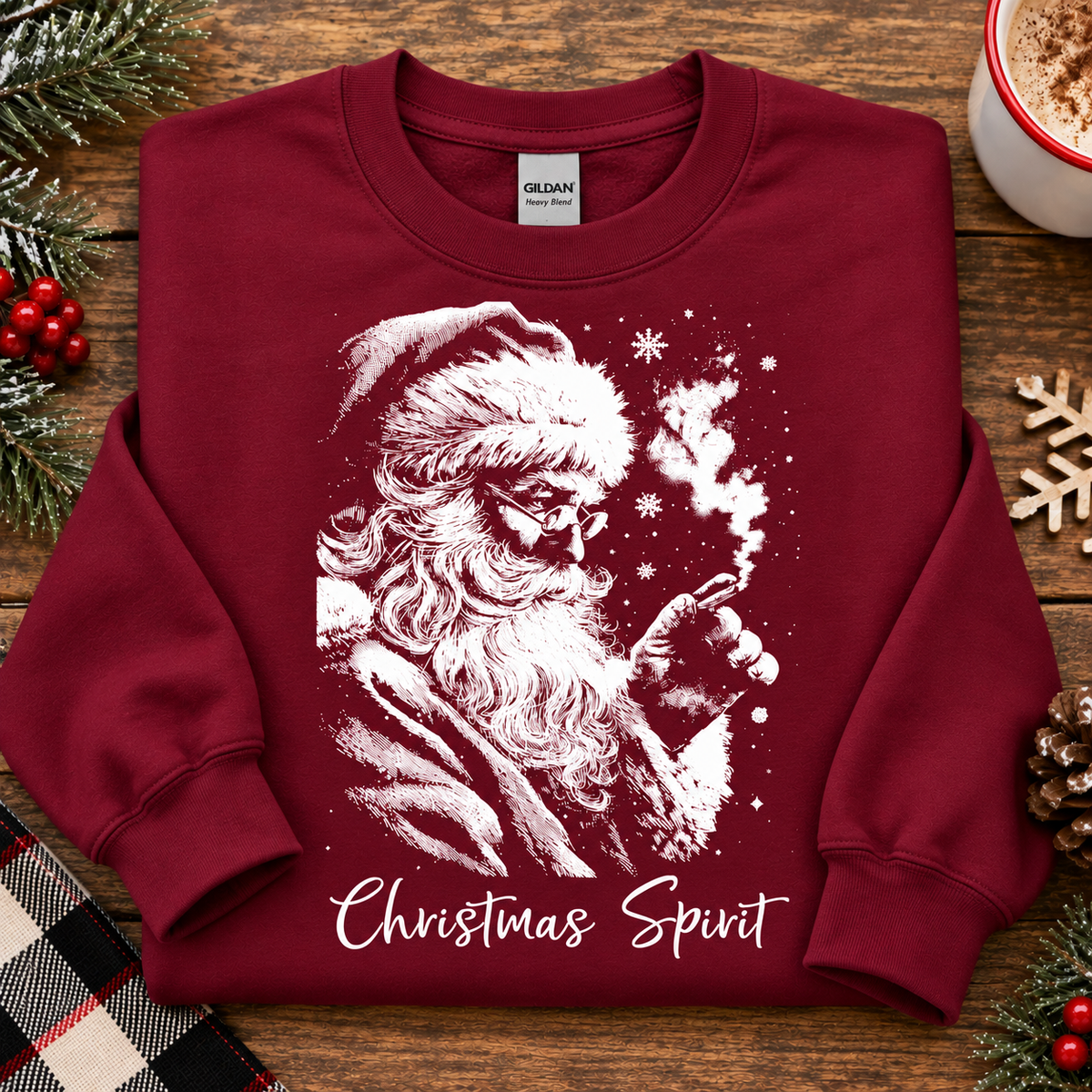 A folded Gildan crewneck sweatshirt in cardinal red, featuring a white graphic of Santa Claus smoking a pipe with snowflakes and the text 'Christmas Spirit' written below. The sweatshirt is styled in a flat-lay on a rustic wooden table surrounded by holiday decorations including pine branches, red berries, a plaid blanket, and a mug of hot cocoa.