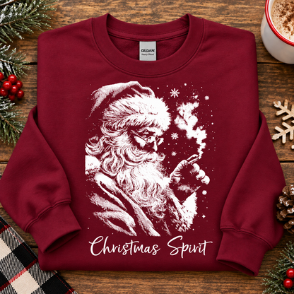 A folded Gildan crewneck sweatshirt in cardinal red, featuring a white graphic of Santa Claus smoking a pipe with snowflakes and the text 'Christmas Spirit' written below. The sweatshirt is styled in a flat-lay on a rustic wooden table surrounded by holiday decorations including pine branches, red berries, a plaid blanket, and a mug of hot cocoa.