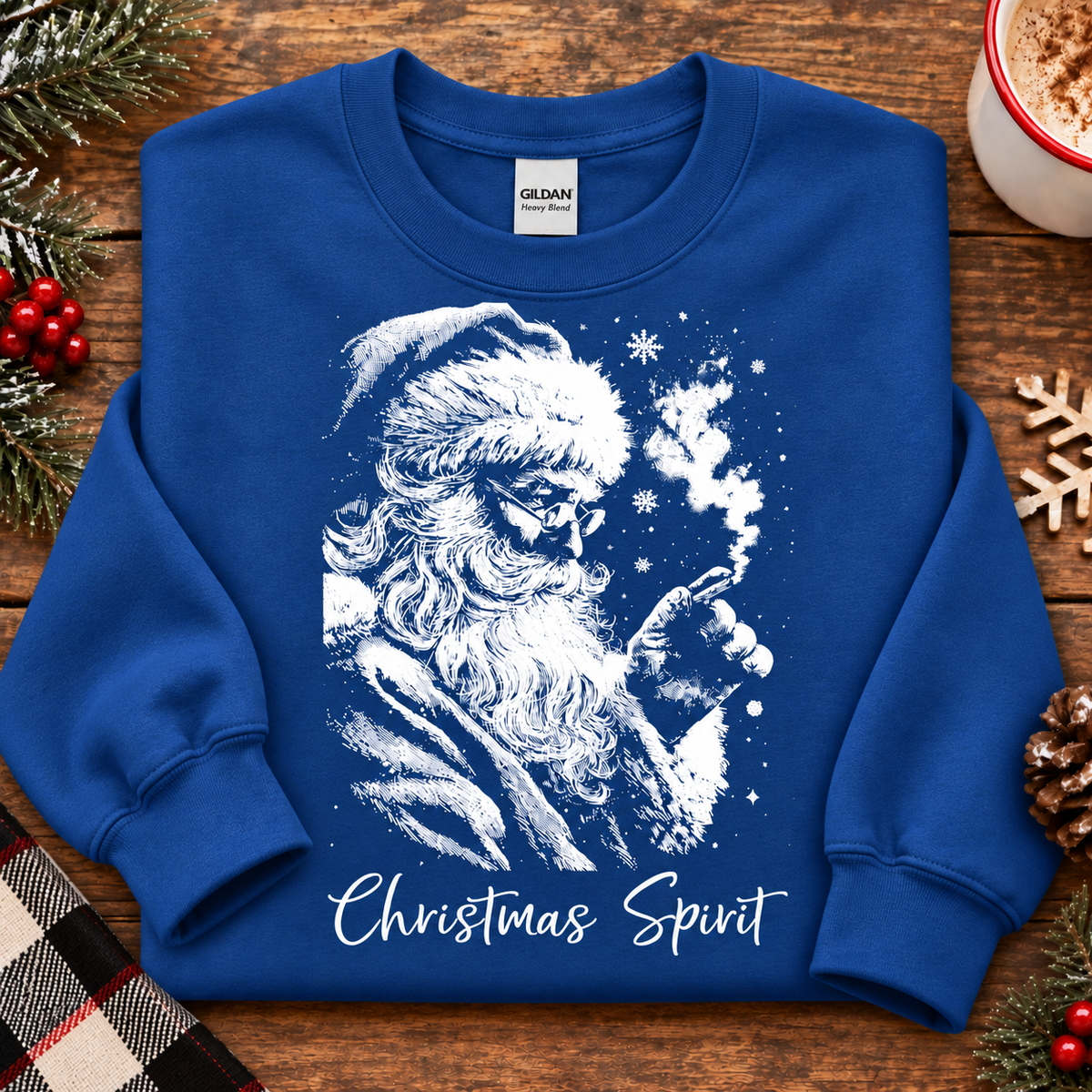 A royal blue crewneck sweatshirt with a white graphic of Santa Claus smoking a pipe, with the text 'Christmas Spirit' written below. The sweatshirt is laid flat on a rustic wooden table surrounded by holiday decorations including pine branches, red berries, a wooden snowflake, a pinecone, and a cup of cocoa.