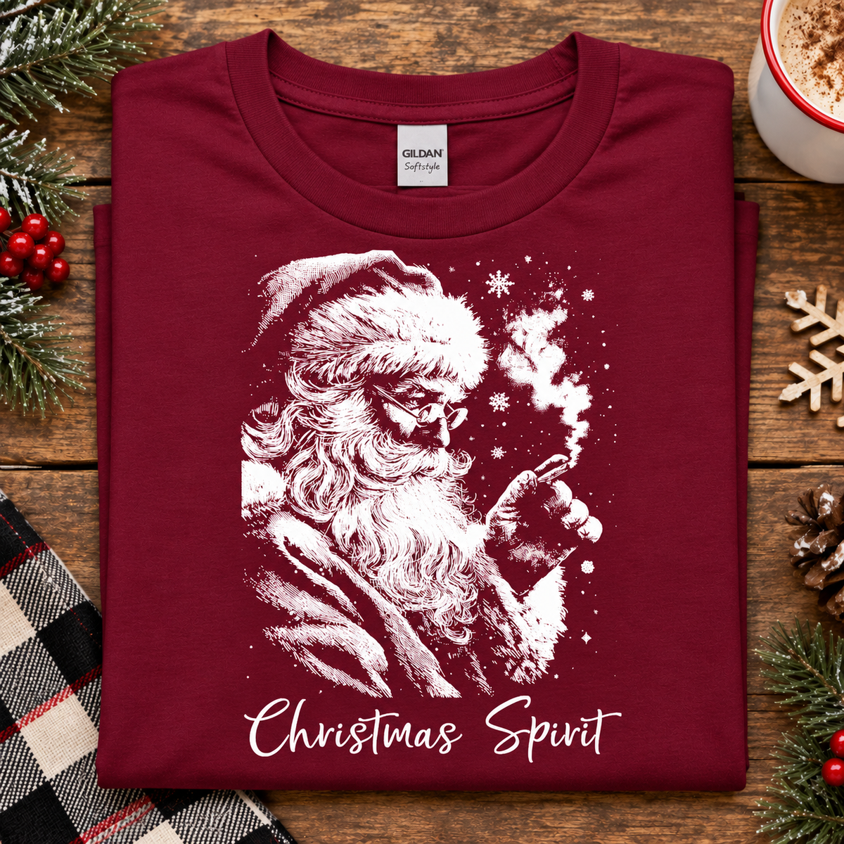 A folded cardinal red crewneck t-shirt with a white graphic of Santa Claus smoking a pipe, with the text 'Christmas Spirit' printed below. The shirt is displayed on a rustic wooden table surrounded by holiday decorations including pine branches, a plaid blanket, and a mug.