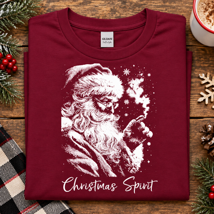 A folded cardinal red crewneck t-shirt with a white graphic of Santa Claus smoking a pipe, with the text 'Christmas Spirit' printed below. The shirt is displayed on a rustic wooden table surrounded by holiday decorations including pine branches, a plaid blanket, and a mug.