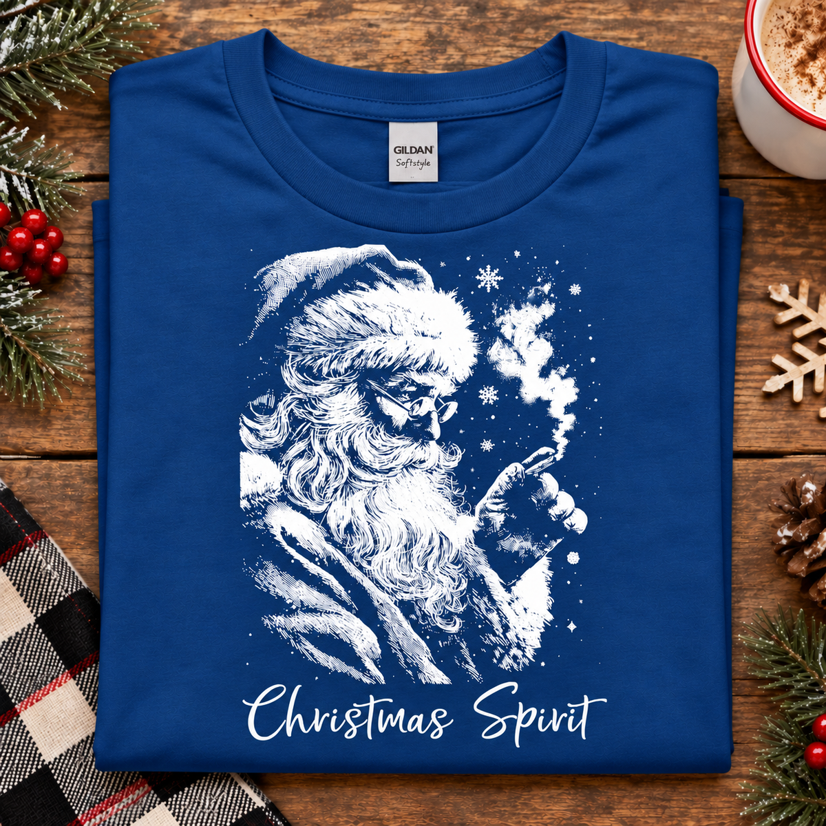 A folded royal blue Gildan Straightforward t-shirt with a white graphic of Santa Claus smoking a pipe, with the text 'Christmas Spirit' printed below. The shirt is displayed on a rustic wooden background with holiday decorations including pine branches, a mug, and a plaid blanket.