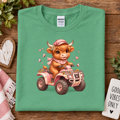 A folded heather irish green Gildan cotton t-shirt with a cute cartoon graphic of a brown cow driving a pink ATV, displayed on a rustic wooden background with decorative props.