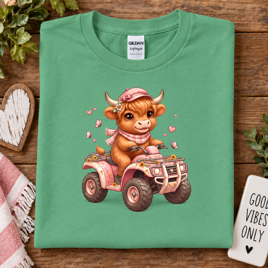 A folded heather irish green Gildan cotton t-shirt with a cute cartoon graphic of a brown cow driving a pink ATV, displayed on a rustic wooden background with decorative props.