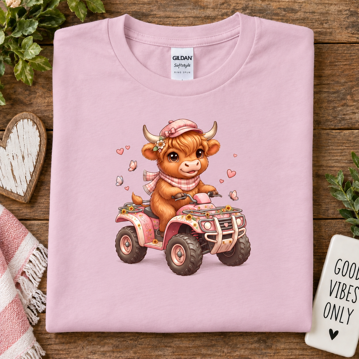 A folded light pink Gildan Straightforward t-shirt is displayed on a rustic wooden background. The shirt features a colorful graphic of a cute brown cow driving a pink ATV, surrounded by small hearts.