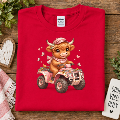 A folded red Gildan cotton t-shirt is displayed on a rustic wooden surface. The front of the shirt features a cute cartoon illustration of a brown cow wearing a pink hat and scarf, riding a pink and orange ATV.