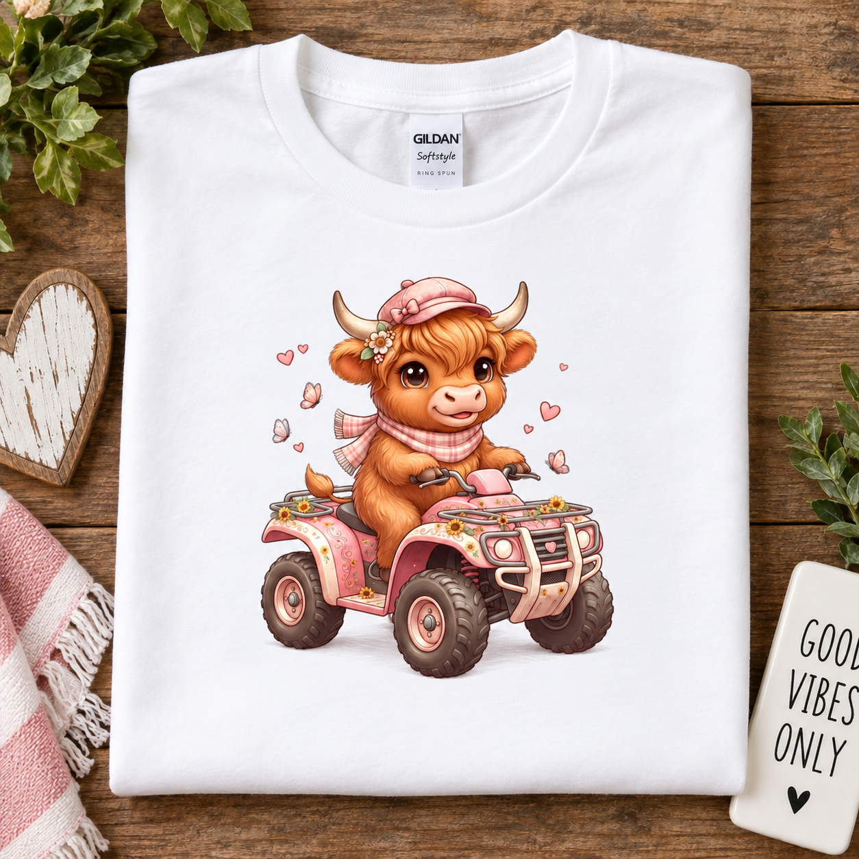 A folded white Gildan Softstyle T-shirt is displayed on a rustic wooden surface, featuring a cute cartoon illustration of a brown cow driving a pink ATV. 