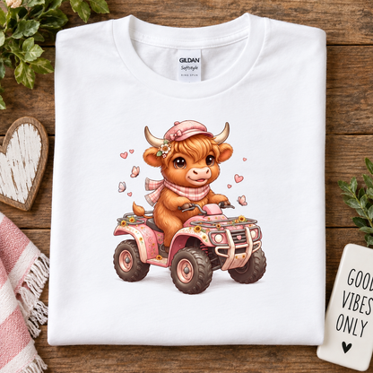 A folded white Gildan Softstyle T-shirt is displayed on a rustic wooden surface, featuring a cute cartoon illustration of a brown cow driving a pink ATV. 