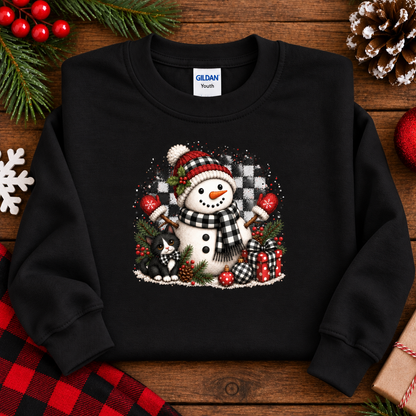 A black crewneck sweatshirt with a Christmas snowman graphic, laid flat on a rustic wooden background with holiday decorations like pine branches, red berries, and a plaid cloth.