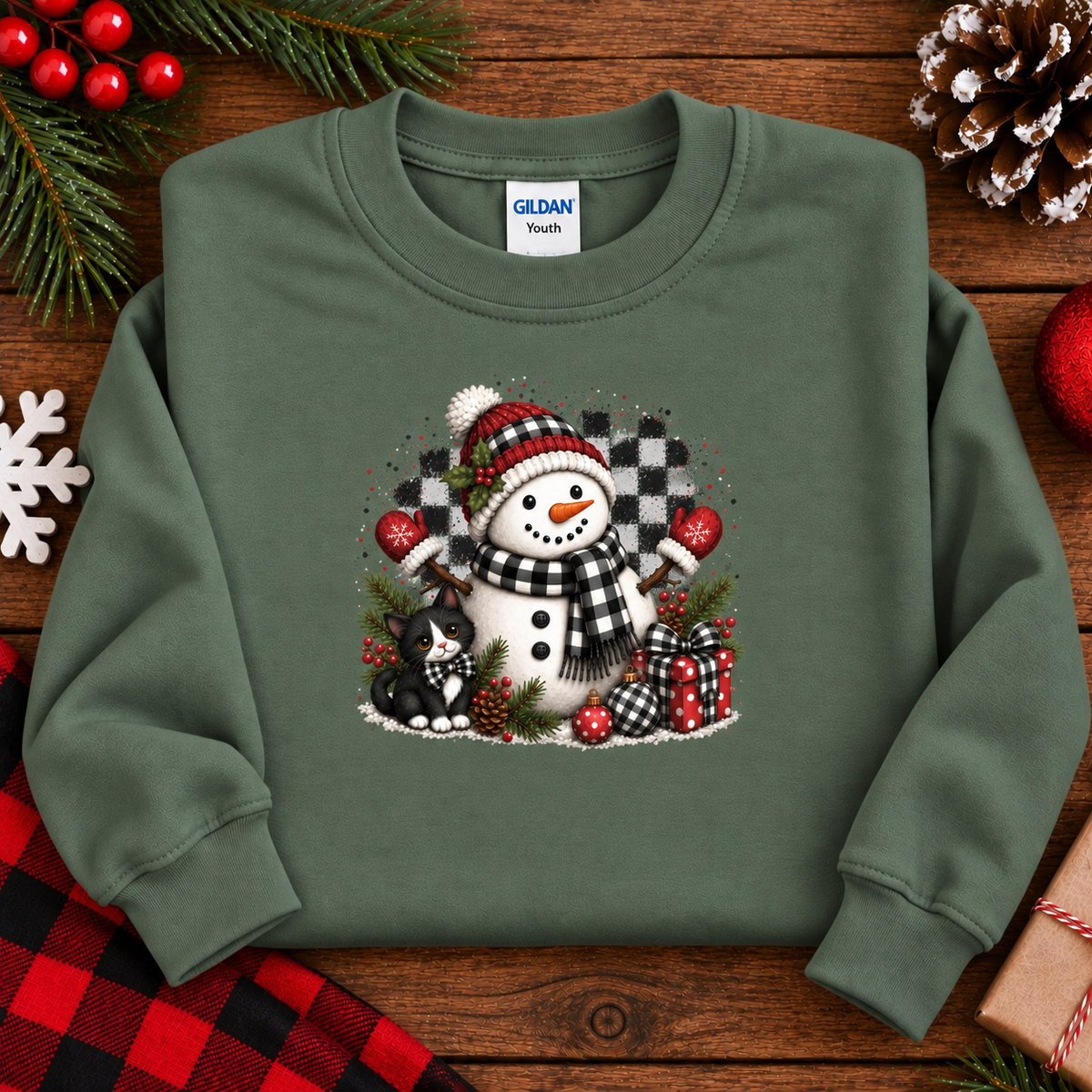 An miliitary green crewneck sweatshirt featuring a festive Christmas graphic of a snowman, a black cat, pinecones, and wrapped gifts, laid flat on a rustic wooden background with holiday decorations.