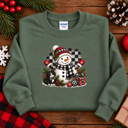 An miliitary green crewneck sweatshirt featuring a festive Christmas graphic of a snowman, a black cat, pinecones, and wrapped gifts, laid flat on a rustic wooden background with holiday decorations.