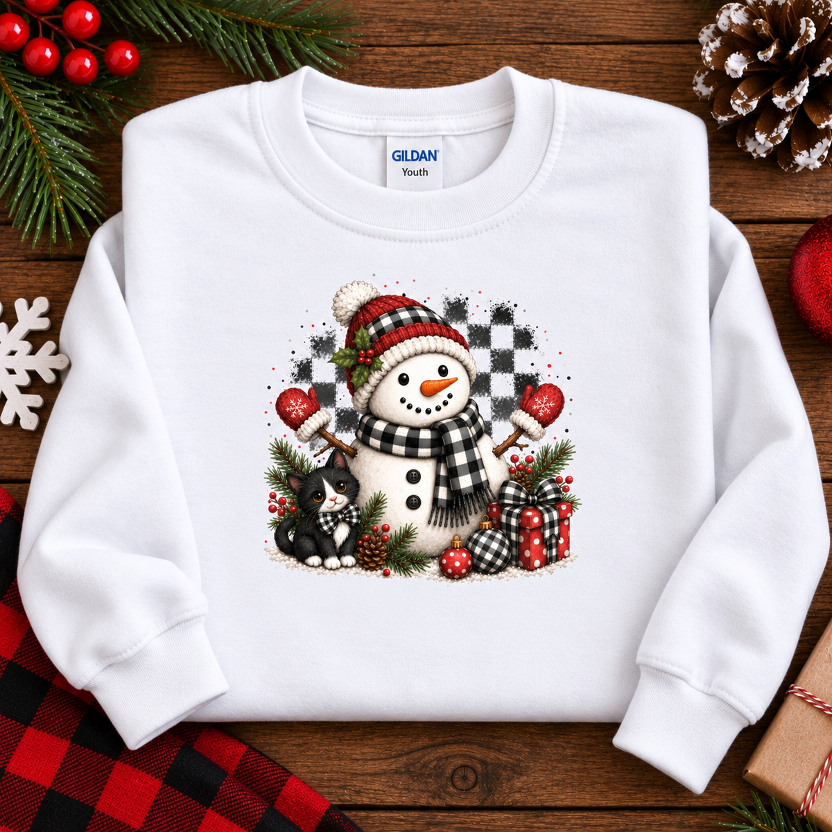 A white Gildan Youth crewneck sweatshirt laid flat on a rustic wooden surface. The front features a colorful graphic of a snowman wearing a red and black checkered hat and scarf, next to a small black cat, with red hearts, pinecones, and wrapped gifts.