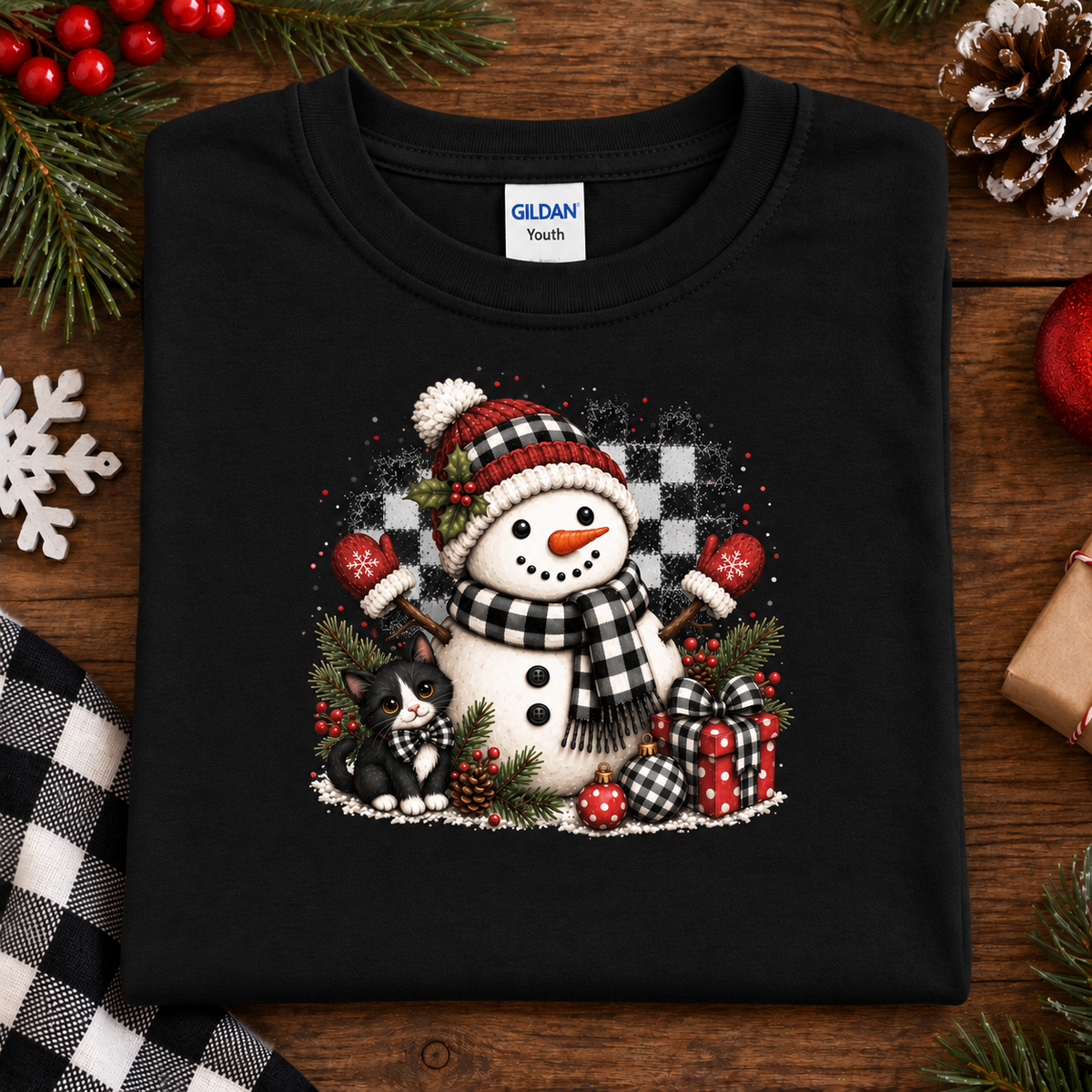 A black Gildan Youth t-shirt folded flat on a rustic wooden surface.The shirt features a festive graphic of a snowman wearing a red and black plaid hat and scarf, holding a pitchfork, with a small black cat and wrapped presents.