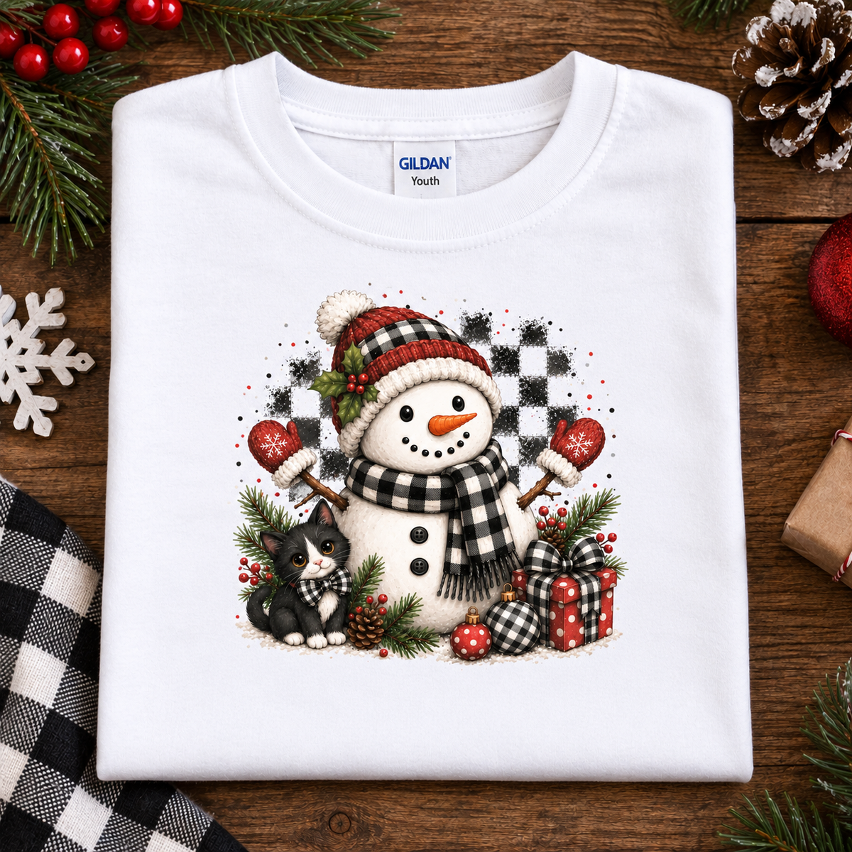 A folded white Gildan Youth t-shirt with a festive Christmas snowman and cat graphic, displayed on a rustic wooden background with holiday decorations.