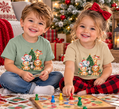 Two children sitting on a white rug in a cozy holiday living room, wearing matching Christmas-themed graphic t-shirts. The boy wears a sage green shirt and the girl wears a beige shirt, both featuring a cartoon animal print of a giraffe, lion, elephant, and mouse around a Christmas tree.