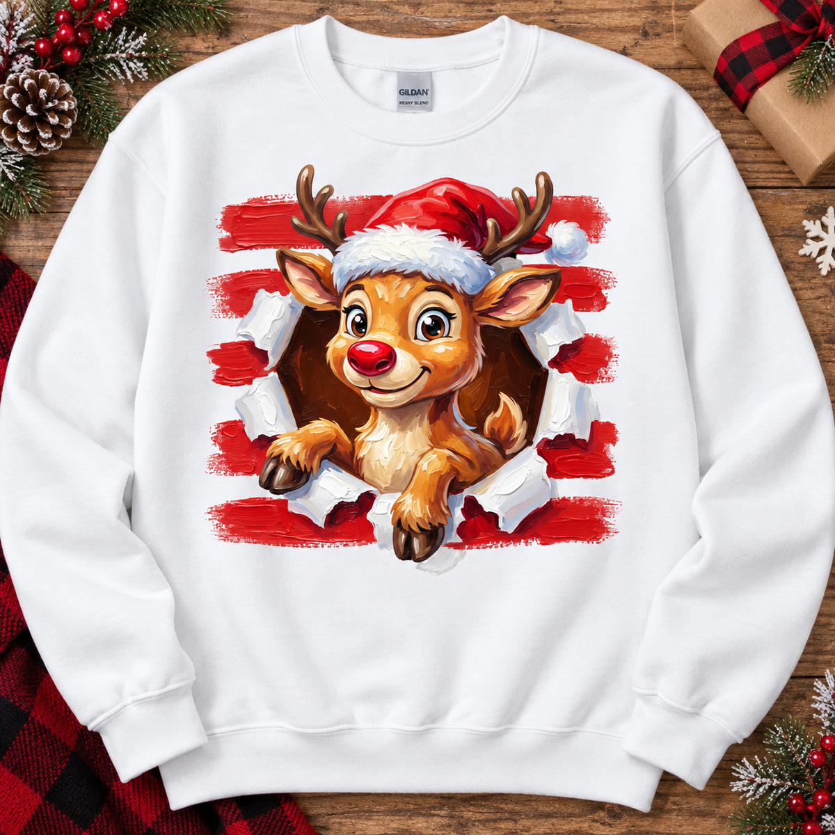 A white crewneck sweatshirt featuring a graphic print of a cute cartoon reindeer wearing a Santa hat, bursting through torn red and white paper. The sweatshirt is displayed flat on a rustic wooden background decorated with holiday elements like pinecones, red berries, and wrapped gifts.