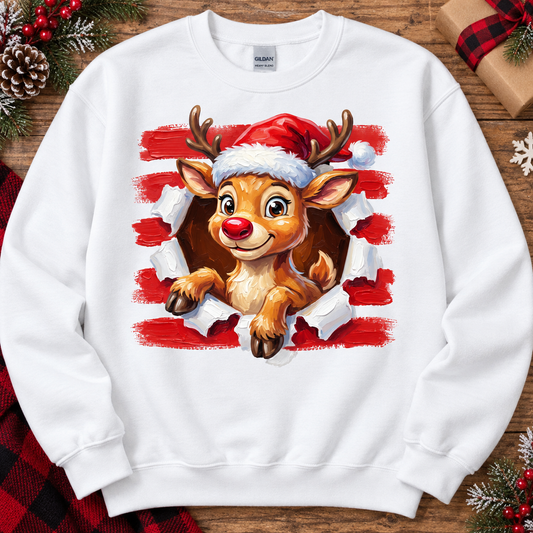 A white crewneck sweatshirt featuring a graphic print of a cute cartoon reindeer wearing a Santa hat, bursting through torn red and white paper. The sweatshirt is displayed flat on a rustic wooden background decorated with holiday elements like pinecones, red berries, and wrapped gifts.