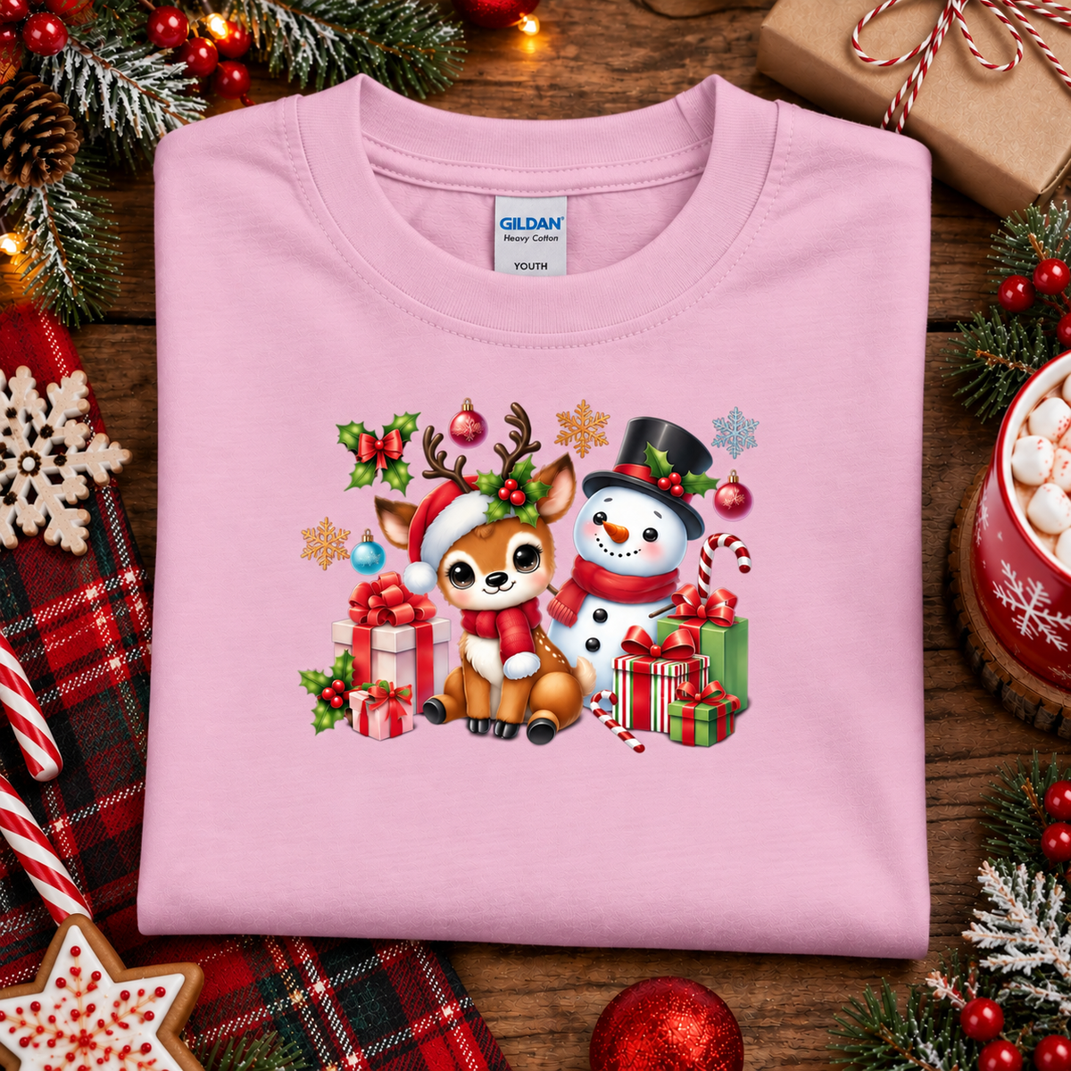 A folded light pink crewneck sweatshirt by Gildan, featuring a colorful Christmas graphic of a deer and a snowman surrounded by gifts. The sweatshirt is styled in a festive flat-lay on a wooden surface with holiday decorations including pine branches, red ornaments, and plaid fabric.