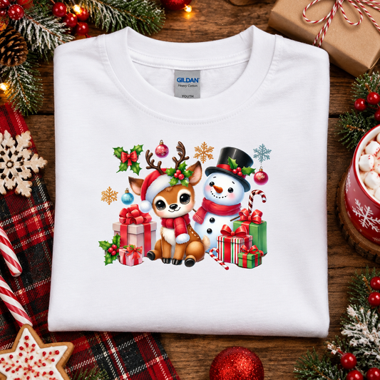 A folded white Gildan T-shirt with a colorful Christmas graphic of a deer and snowman surrounded by gifts, displayed on a rustic wooden surface with holiday decorations.