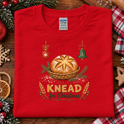 A folded red Gildan Softstyle T-shirt with a Christmas-themed bread graphic and the text 'ALL I KNEAD for Christmas,' styled on a wooden surface with holiday decorations and plaid fabric around it.