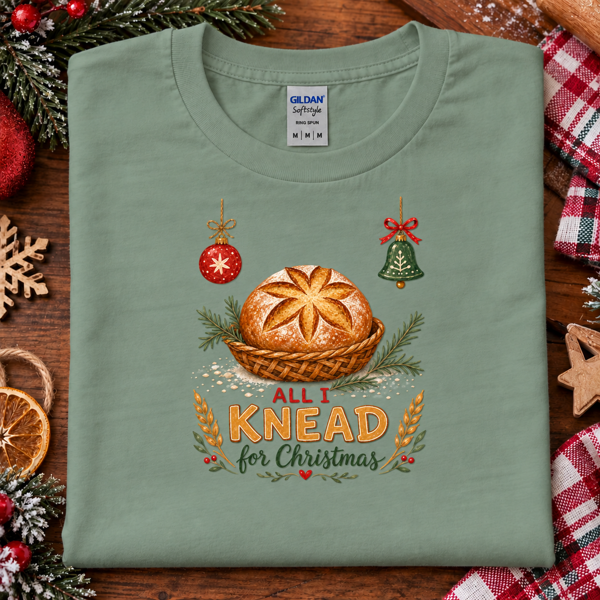 A sage green Gildan Softstyle features a graphic of a golden-brown loaf of bread in a basket, flanked by a red ornament and a green bell, with the text 'ALL I KNEAD for Christmas' written below.