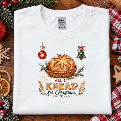 A folded white Gildan Softstyle T-shirt is displayed on a rustic wooden surface with Christmas-themed decorative props, including evergreen branches, a red ornament, a green bell, a dried orange slice, and a plaid fabric. The shirt features a graphic of a baked pie in a basket with the text 'ALL I KNEAD for Christmas'.