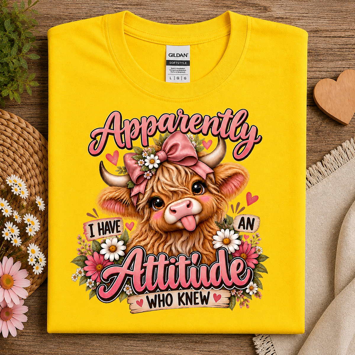 A folded daisy colored Gildan Ultra-Cotton T-shirt is displayed on a wooden surface with decorative props including flowers, a woven hat, and a beige cloth. The shirt features a colorful graphic of a fluffy Highland cow wearing a pink bow and surrounded by flowers, with the text 'Apparently I HAVE AN Attitude WHO KNEW'. 