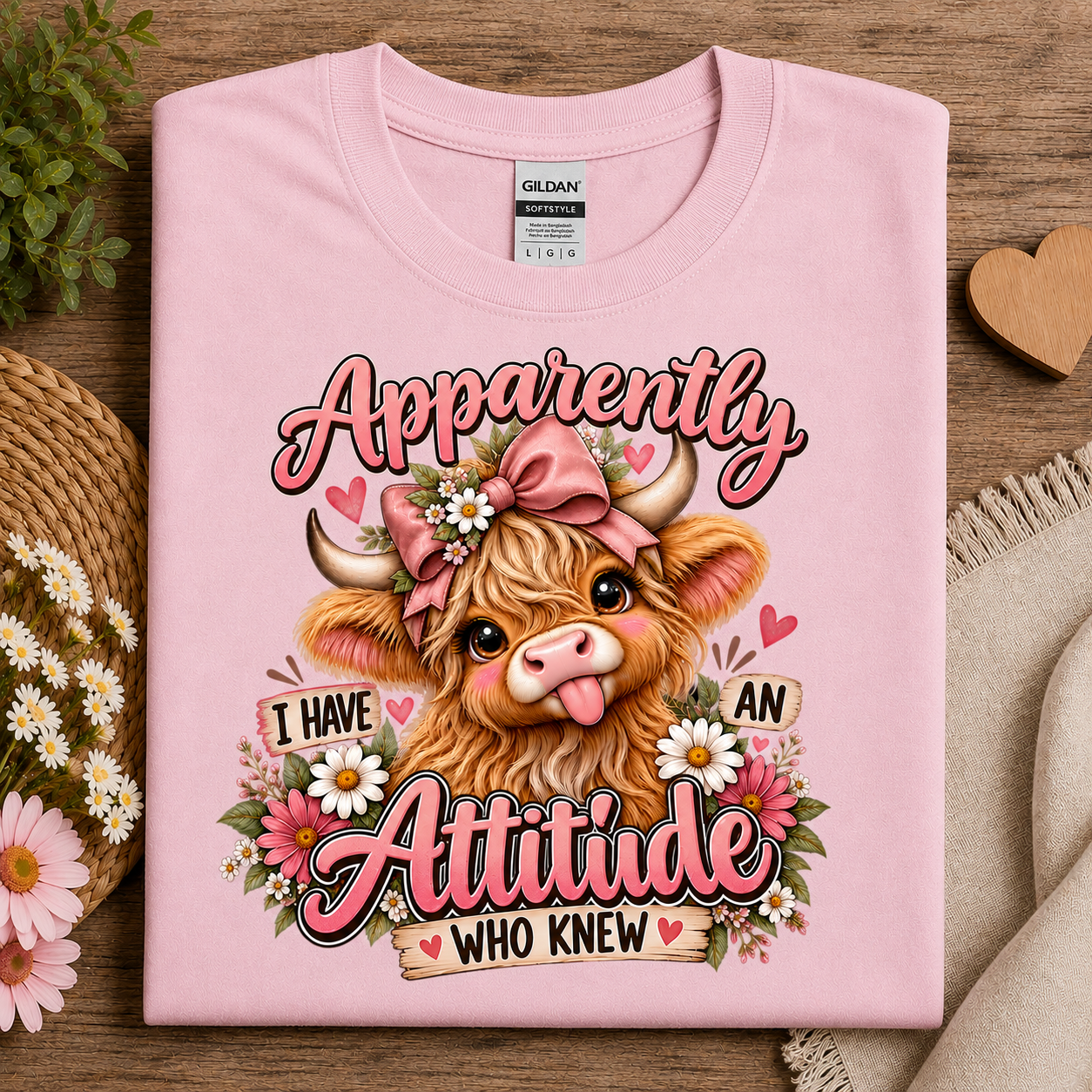 A folded light pink Gildan t-shirt with a graphic print of a highland cow wearing a pink bow and surrounded by flowers, with the text 'Apparently I HAVE AN Attitude WHO KNEW'. The shirt is styled on a wooden surface with decorative props including daisies, a straw hat, and a beige throw blanket.