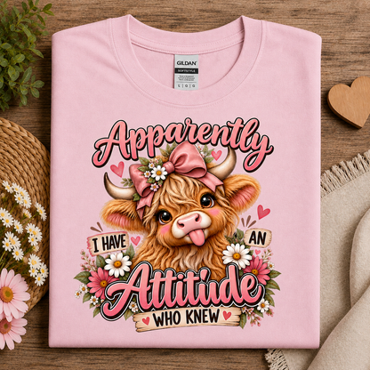 A folded light pink Gildan t-shirt with a graphic print of a highland cow wearing a pink bow and surrounded by flowers, with the text 'Apparently I HAVE AN Attitude WHO KNEW'. The shirt is styled on a wooden surface with decorative props including daisies, a straw hat, and a beige throw blanket.