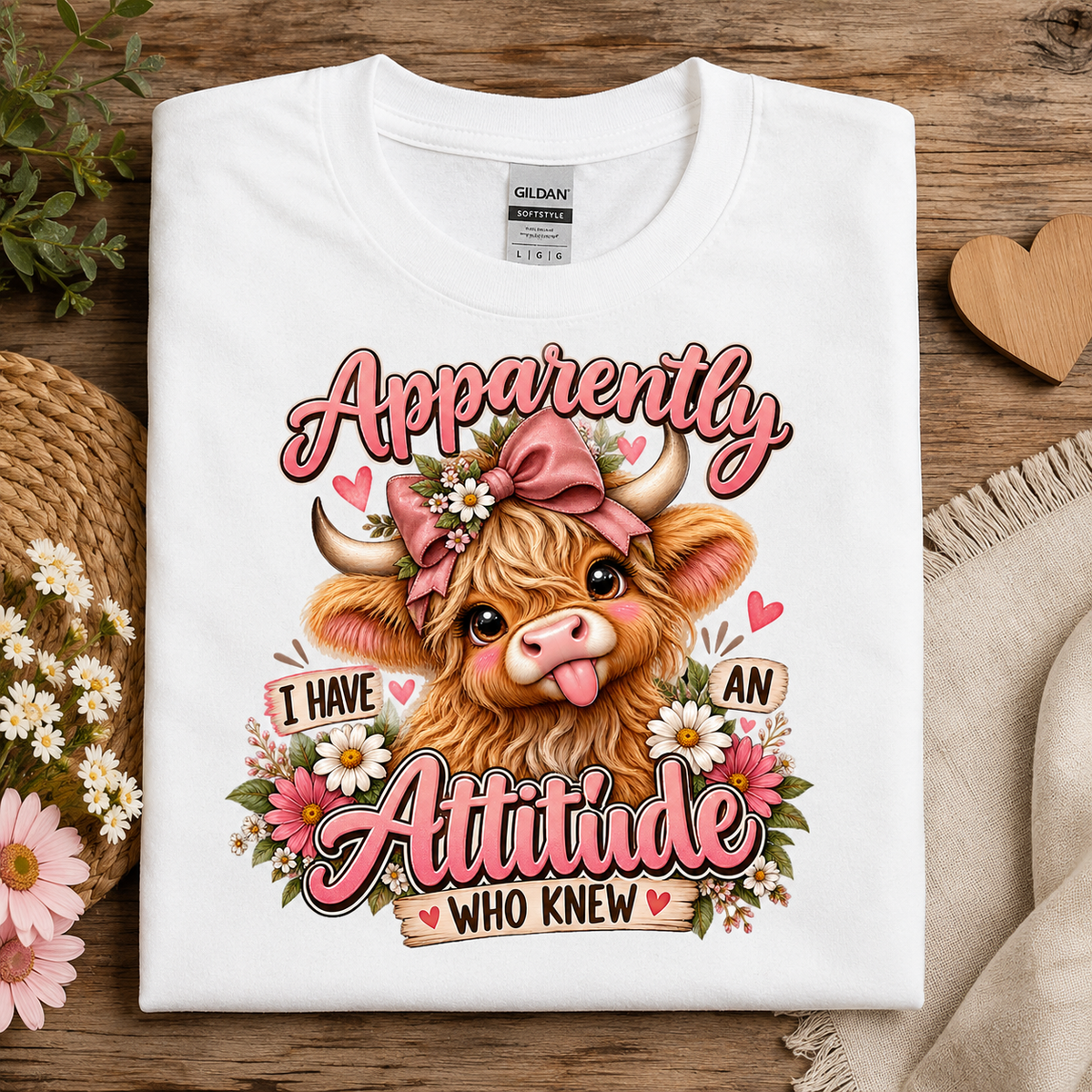 A folded white Gildan Comfort Colors T-shirt is displayed on a rustic wooden surface with decorative props including flowers, a woven mat, and a wooden heart. The shirt features a colorful graphic of a fluffy brown Scottish Highland cow wearing a pink bow and floral crown, surrounded by text that reads, 'Apparently I Have An Attitude Who Knew'.