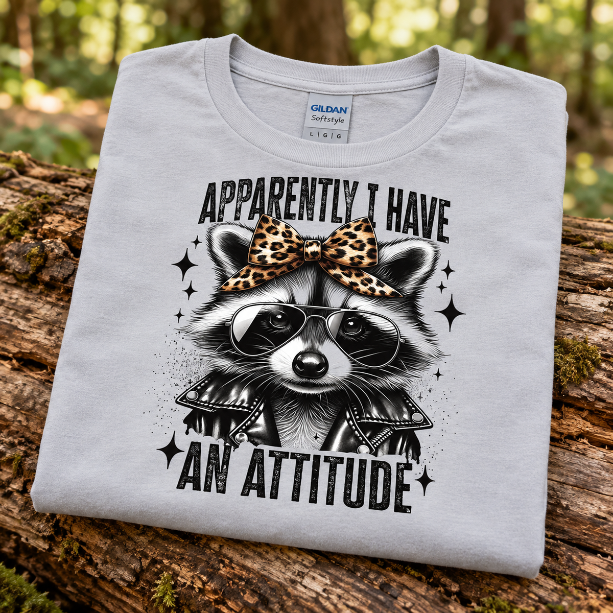 A folded light gray Gildan Comfort t-shirt is displayed on a rustic log in a forest setting. The shirt features a graphic of a raccoon wearing a leopard-print bow, black sunglasses, and a black leather jacket, with the text 'APPARENTLY I HAVE AN ATTITUDE'.