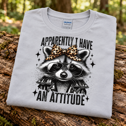 A folded light gray Gildan Comfort t-shirt is displayed on a rustic log in a forest setting. The shirt features a graphic of a raccoon wearing a leopard-print bow, black sunglasses, and a black leather jacket, with the text 'APPARENTLY I HAVE AN ATTITUDE'.