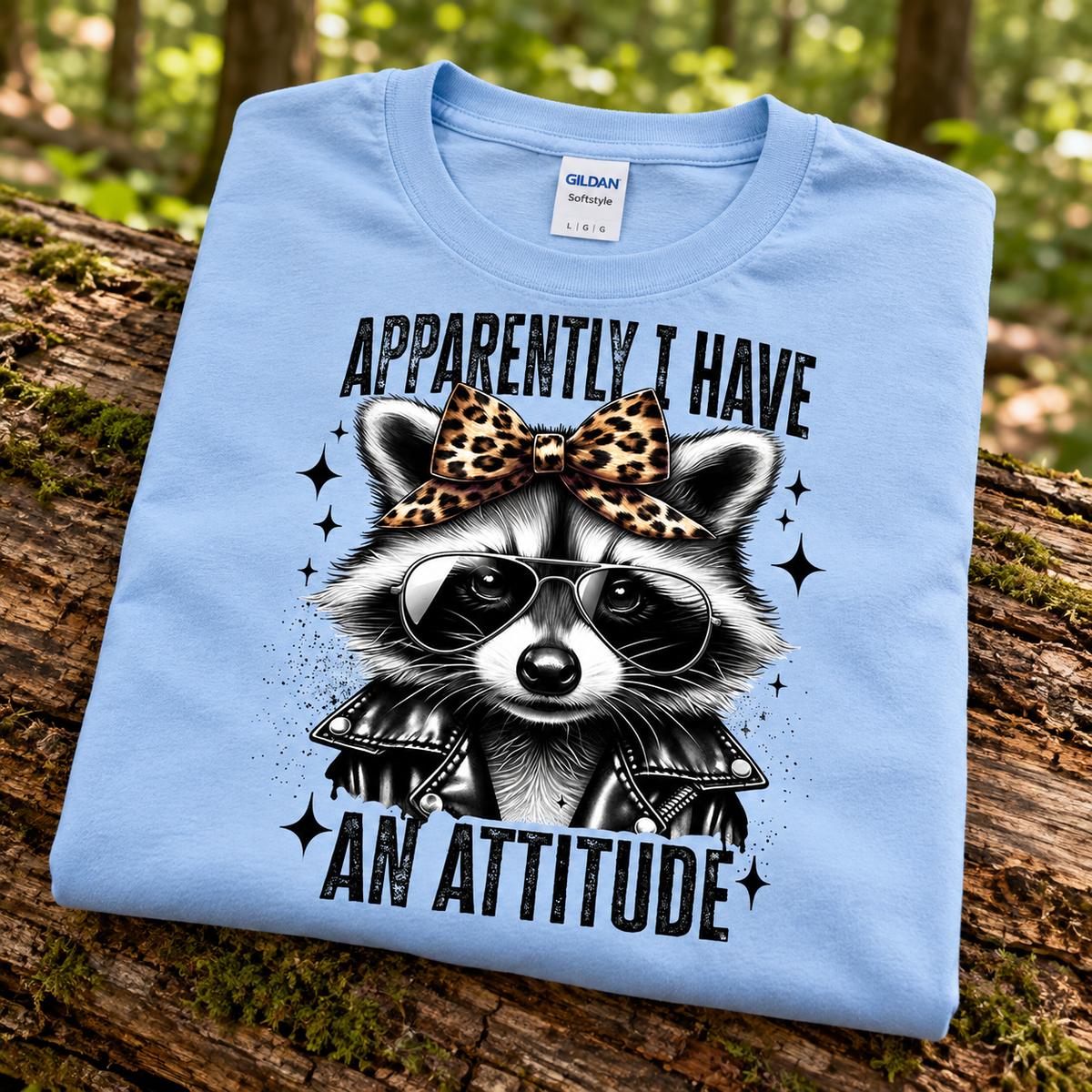 A folded light blue Gildan Blue t-shirt is displayed on a mossy log in a forest setting. The shirt features a graphic of a raccoon wearing a leopard-print bow, black sunglasses, and a black leather jacket, with the text 'APPARENTLY I HAVE AN ATTITUDE' in a black retro font.