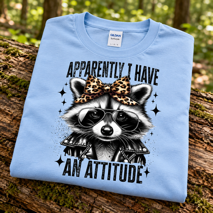 A folded light blue Gildan Blue t-shirt is displayed on a mossy log in a forest setting. The shirt features a graphic of a raccoon wearing a leopard-print bow, black sunglasses, and a black leather jacket, with the text 'APPARENTLY I HAVE AN ATTITUDE' in a black retro font.