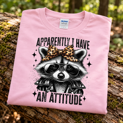 A folded light pink Gildan t-shirt is displayed on a mossy log outdoors. The shirt features a graphic of a raccoon wearing sunglasses and a leopard-print bow, with the text 'APPARENTLY I HAVE AN ATTITUDE'.